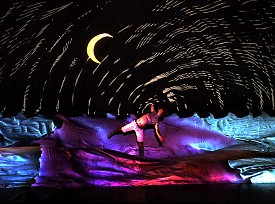 A male performer balancing on one foot in a colourful sea of cloth underneath the night sky