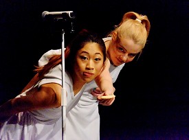 Two dancers in white standing behind a microphone stand looking at a cookie