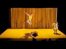 A male dancer in yellow jumping in midair with both arms outstretched