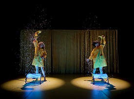 Two dancers in yellow raincoats pouring confetti out of yellow boots above two fans
