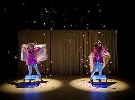 Two dancers in pink standing behind two fans with pink confetti blowing in the air