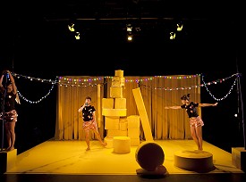 Three dancers on a yellow stage with strings of colourful hanging lights