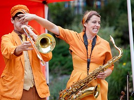 A musician holding a saxophone pinching the nose of another musician playing the trombone